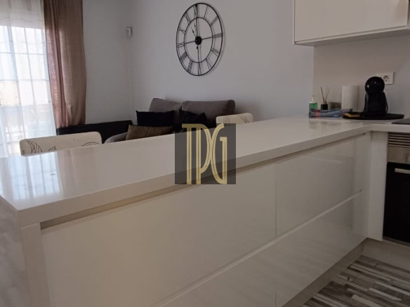 Bright modern kitchen with a white island, warm pendant lights, and a clock on the wall; living area with a sofa visible beyond.