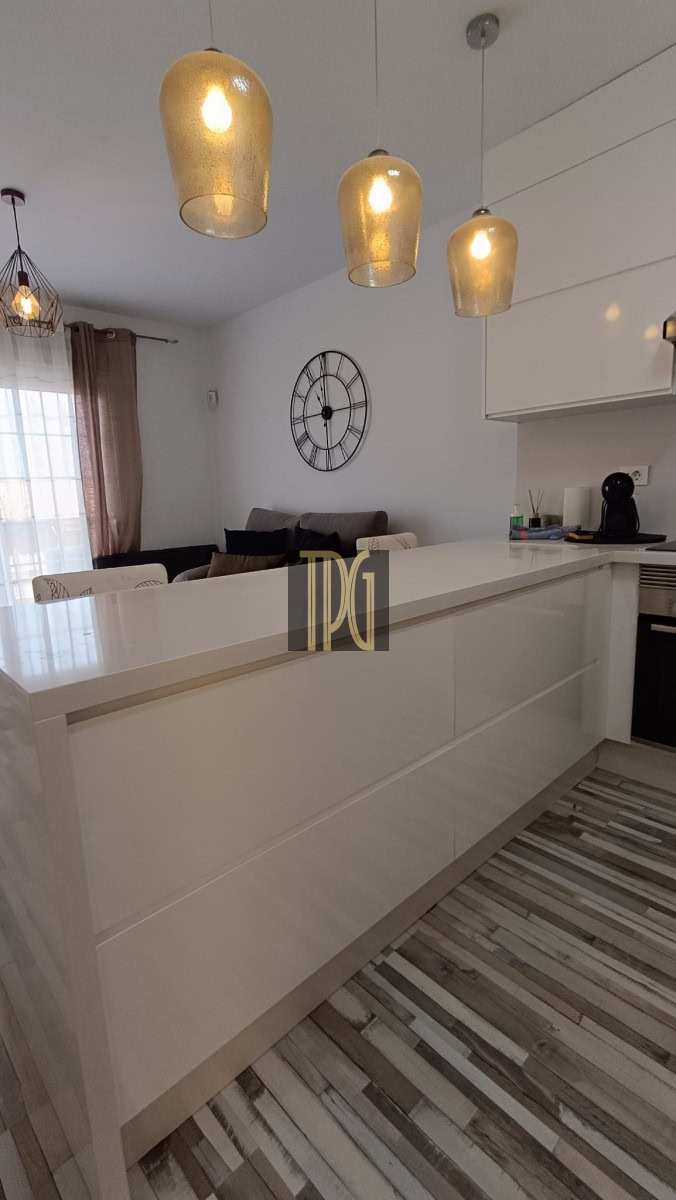 Bright modern kitchen with a white island, warm pendant lights, and a clock on the wall; living area with a sofa visible beyond.