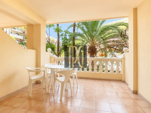 Sunny patio: beige-walled balcony with a white round table and four white chairs, tiled floor, and palm trees beyond the railing.