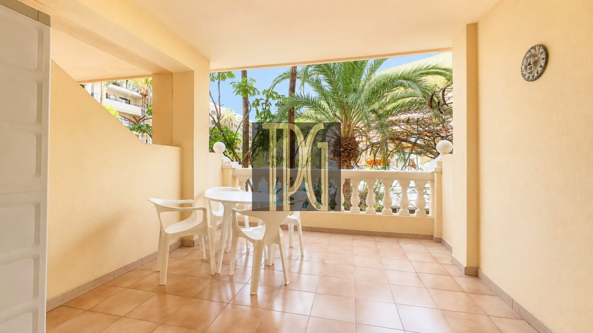 Sunny patio: beige-walled balcony with a white round table and four white chairs, tiled floor, and palm trees beyond the railing.