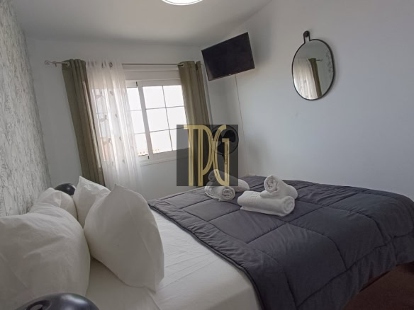 Bedroom with white bedding, gray quilt, and folded towels on the bed; wallpapered accent wall on the left and a window with sheer curtains.