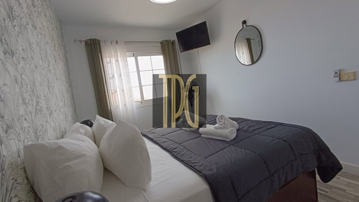 Bedroom with white bedding, gray quilt, and folded towels on the bed; wallpapered accent wall on the left and a window with sheer curtains.