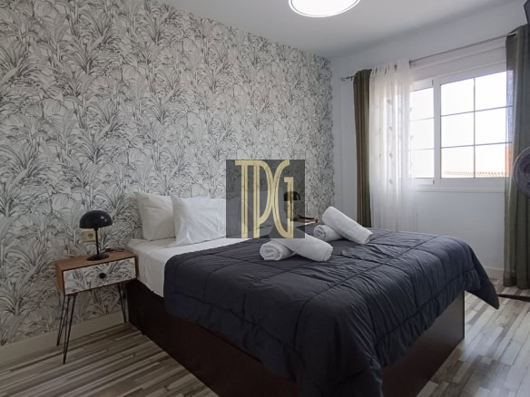 Bedroom with palm-pattern wallpaper, a queen bed with dark gray comforter and white pillows, towels folded on top, and a bedside lamp on a small wooden table.