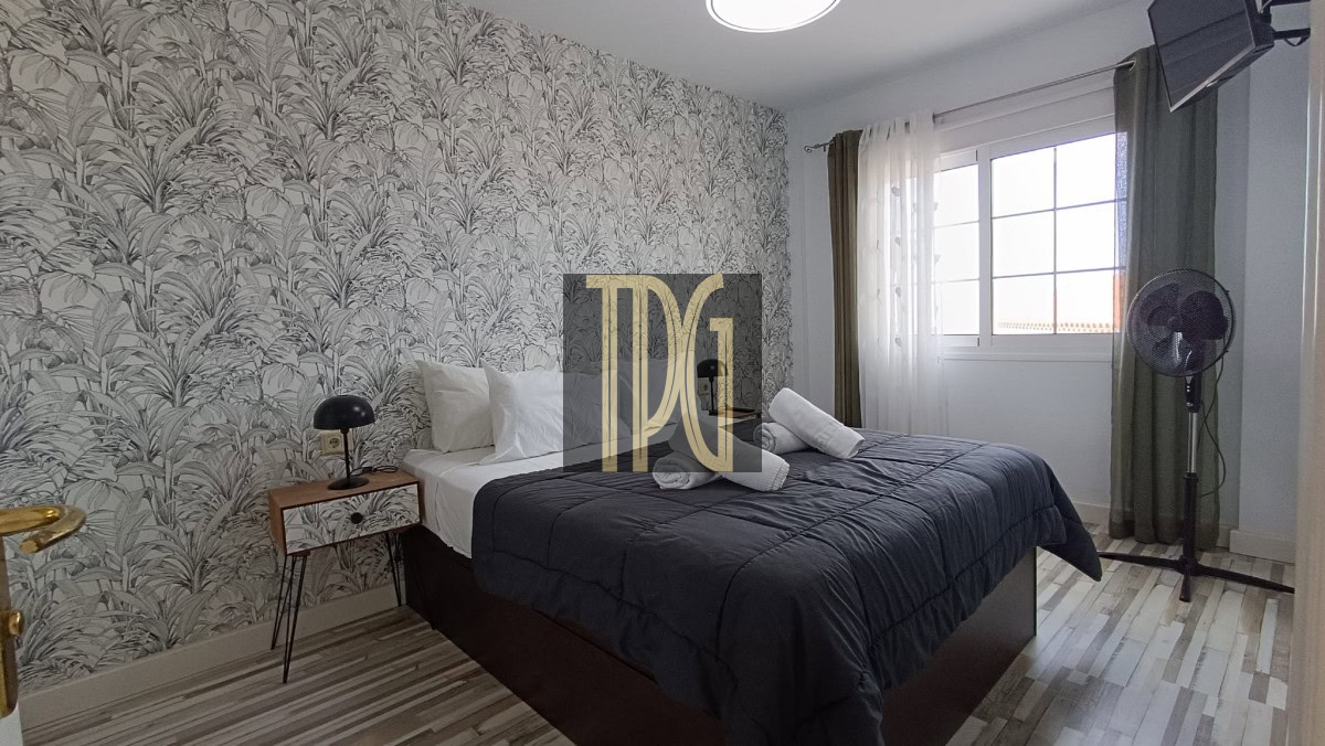 Bedroom with palm-pattern wallpaper, a queen bed with dark gray comforter and white pillows, towels folded on top, and a bedside lamp on a small wooden table.