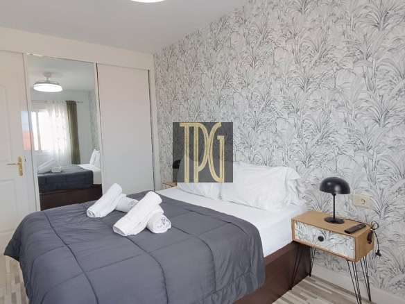 Double bed in a modern bedroom with a dark gray quilt, white pillows, and three rolled towels on the bed; tropical-pattern wallpaper, a small wooden nightstand with a black lamp, and a mirrored wardrobe across from the bed.