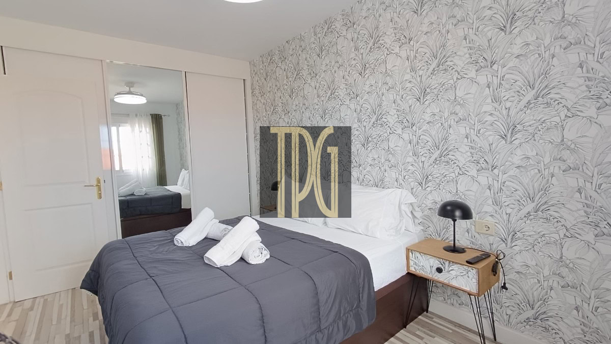 Double bed in a modern bedroom with a dark gray quilt, white pillows, and three rolled towels on the bed; tropical-pattern wallpaper, a small wooden nightstand with a black lamp, and a mirrored wardrobe across from the bed.