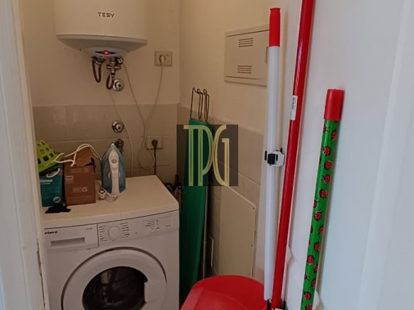 Laundry closet with a front-loading washing machine beneath a tall white water heater; cleaning tools (mop, broom) and a red dustpan nearby.
