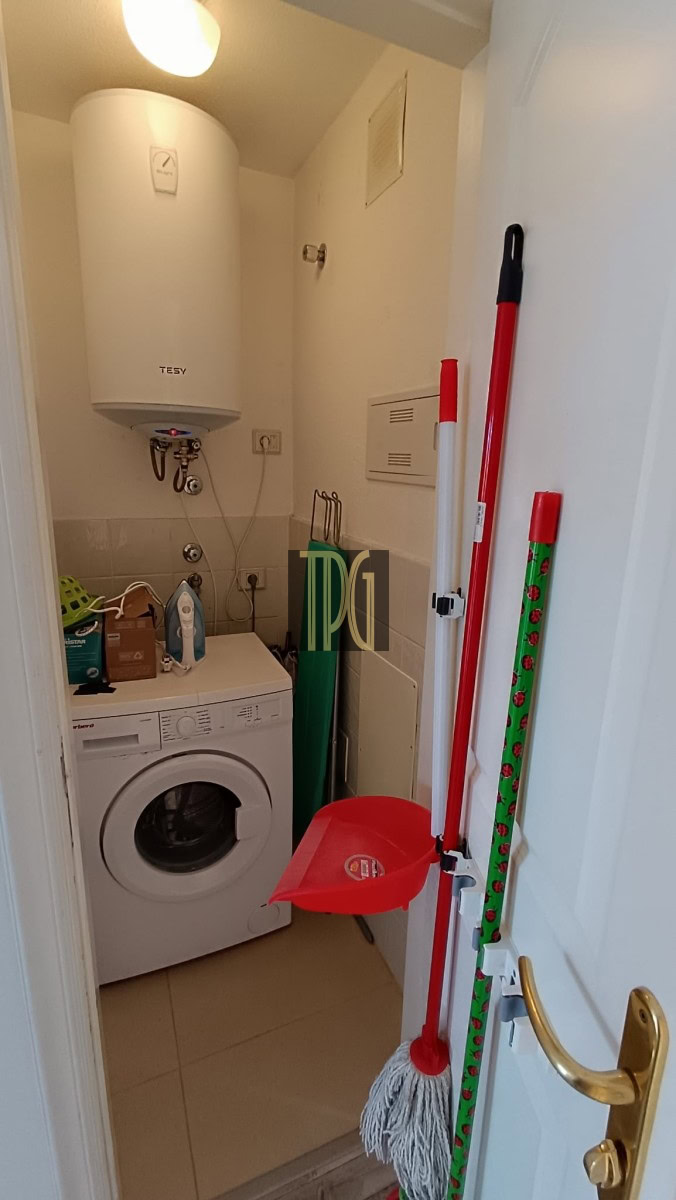 Laundry closet with a front-loading washing machine beneath a tall white water heater; cleaning tools (mop, broom) and a red dustpan nearby.
