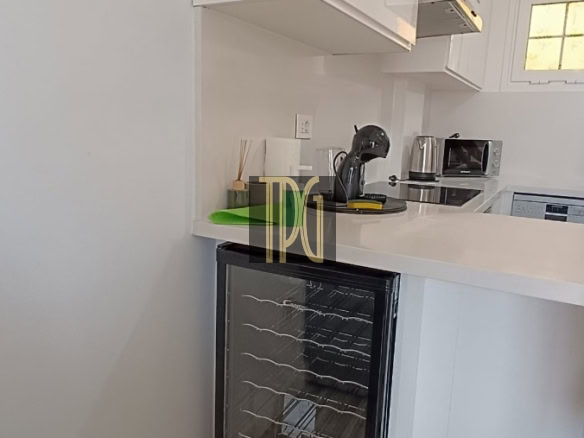 Compact white kitchen with a glass-front wine cooler under the counter, white cabinets, and a countertop with a coffee maker, kettle, and microwave near a small window.