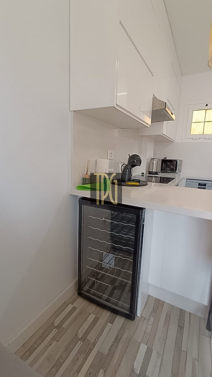 Compact white kitchen with a glass-front wine cooler under the counter, white cabinets, and a countertop with a coffee maker, kettle, and microwave near a small window.