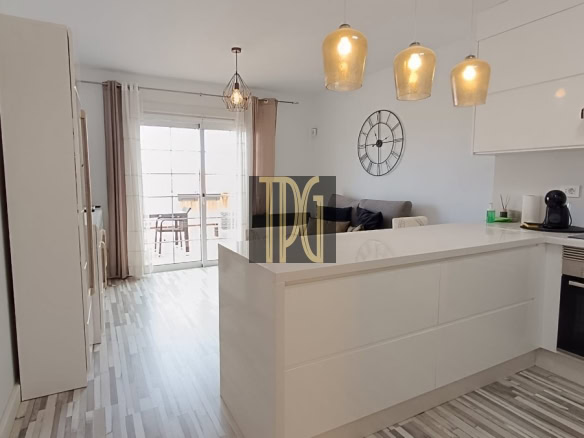 Bright white modern kitchen with a large island, three beige pendant lights, and a wall clock; sliding glass doors to a balcony in the background.