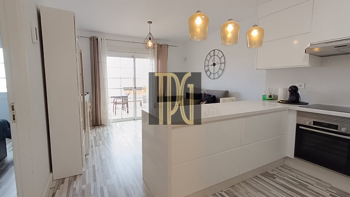 Bright white modern kitchen with a large island, three beige pendant lights, and a wall clock; sliding glass doors to a balcony in the background.