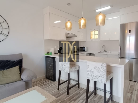 Open-plan living area with a white kitchen island, two upholstered bar stools, a stainless steel fridge, and a large wall clock above a gray sofa.