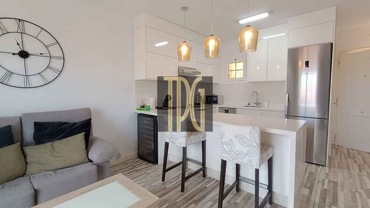 Open-plan living area with a white kitchen island, two upholstered bar stools, a stainless steel fridge, and a large wall clock above a gray sofa.