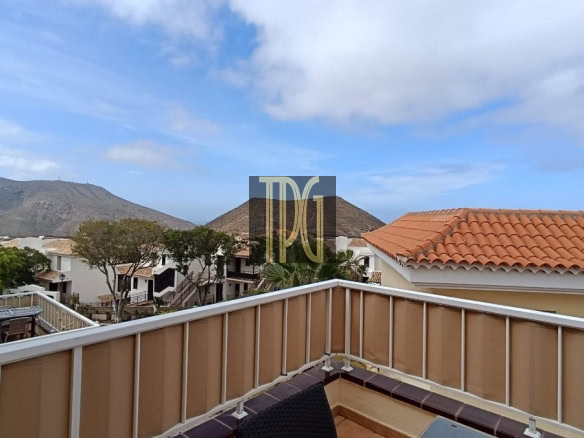 Balcony railing with a dark wicker chair on a tiled terrace overlooking white houses, trees, and distant hills under a blue sky.