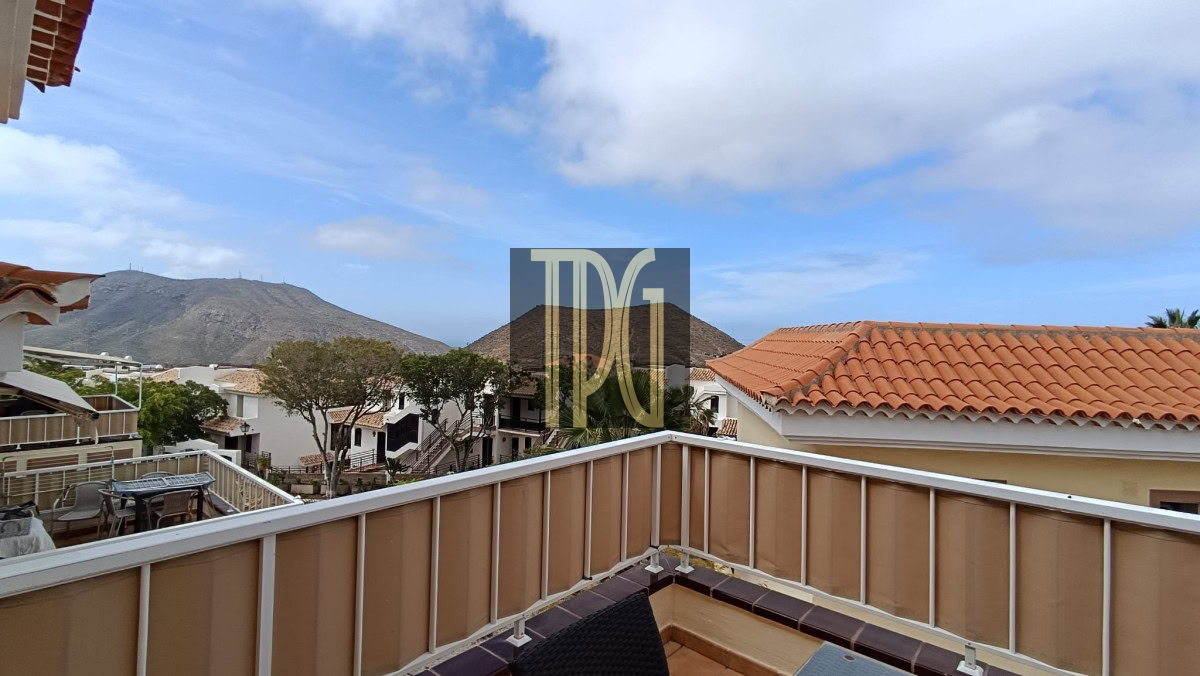 Balcony railing with a dark wicker chair on a tiled terrace overlooking white houses, trees, and distant hills under a blue sky.