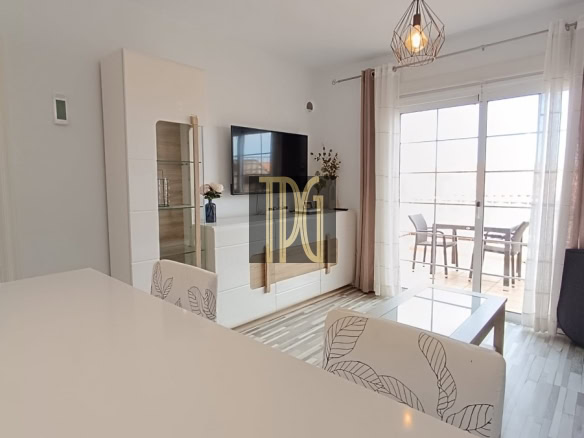 Bright, modern living room with white furniture, a glass coffee table, wall-mounted TV, and a sliding glass door to a balcony.