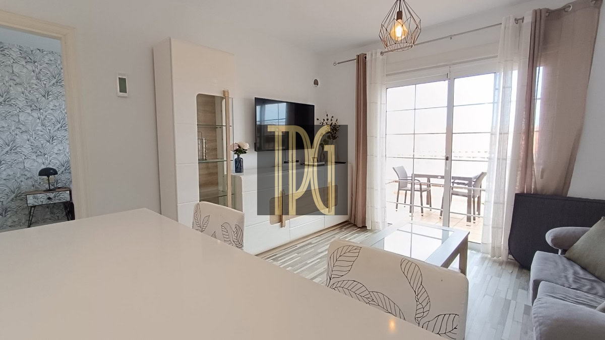 Bright, modern living room with white furniture, a glass coffee table, wall-mounted TV, and a sliding glass door to a balcony.