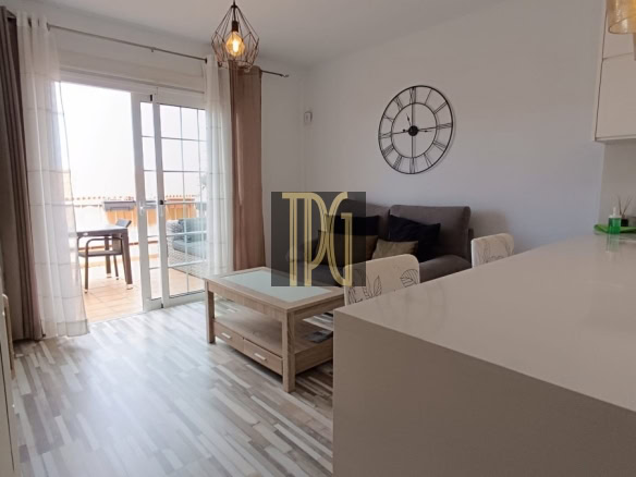 Living room with a gray sofa, wooden coffee table, and a large black wall clock; sliding glass doors to a balcony in the background.