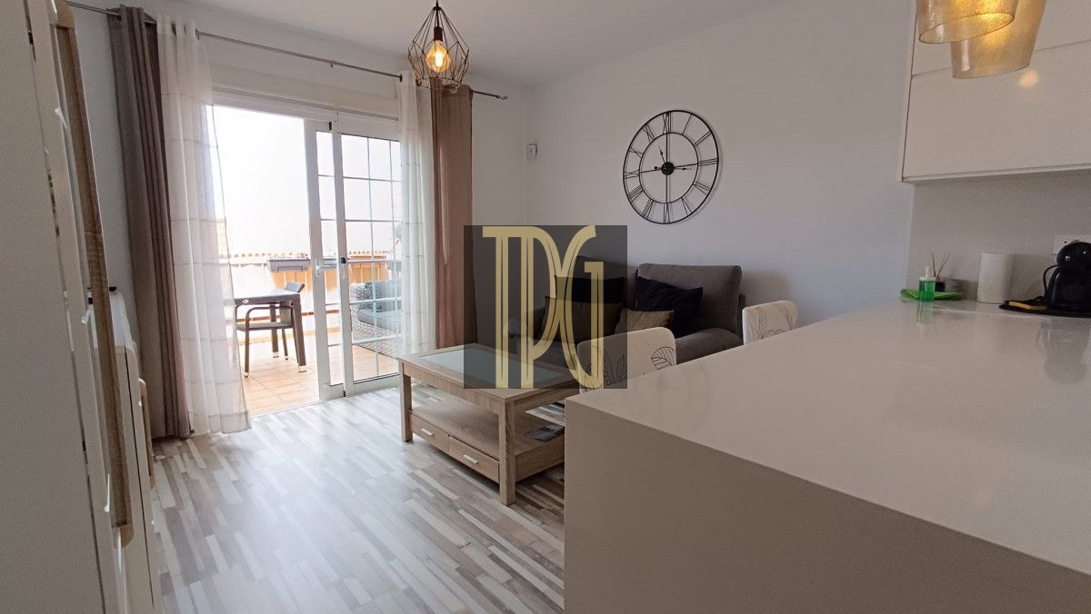 Living room with a gray sofa, wooden coffee table, and a large black wall clock; sliding glass doors to a balcony in the background.
