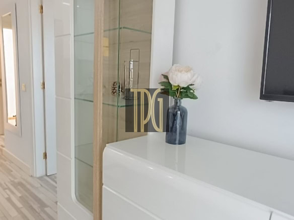 White modern cabinet and glass display with wooden handles; vase of white flowers on top and a wall-mounted TV nearby.
