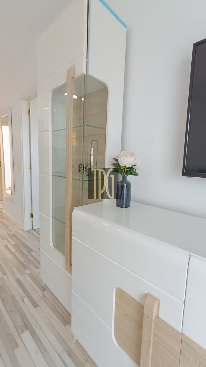 White modern cabinet and glass display with wooden handles; vase of white flowers on top and a wall-mounted TV nearby.