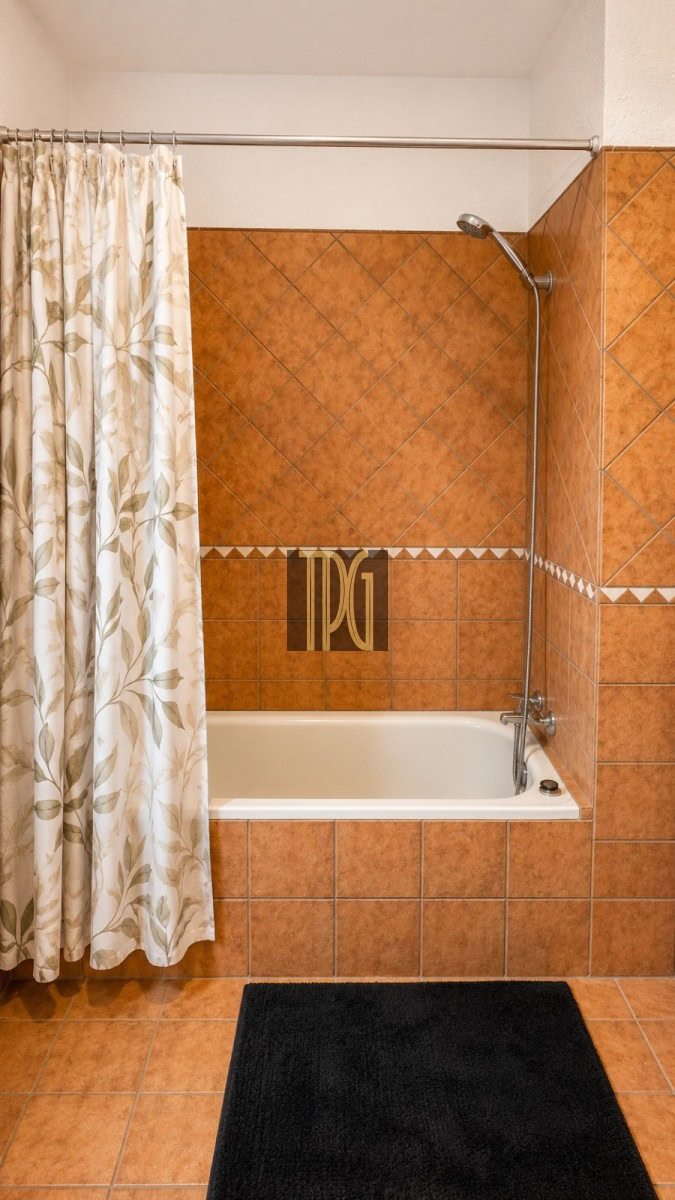 Bathroom with a white tub, orange-brown tiled surround, and a leaf-pattern shower curtain on the left; a dark rug on the floor.
