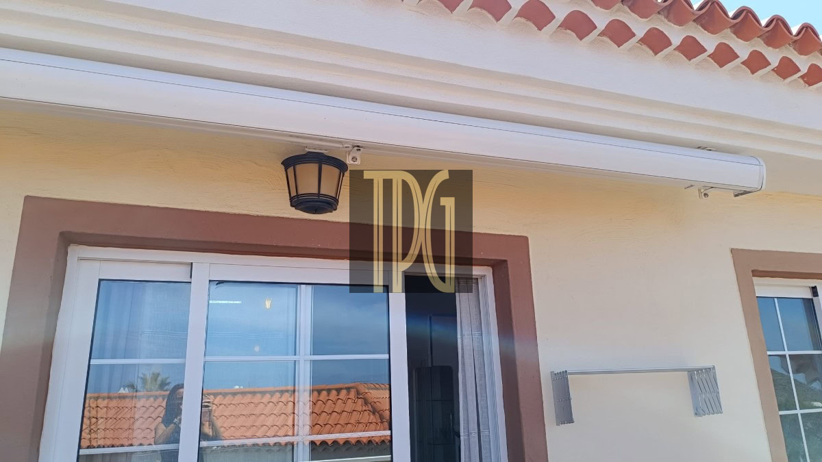 White retractable awning over a glass door on a beige stucco wall with brown trim and a black lantern-style light above.