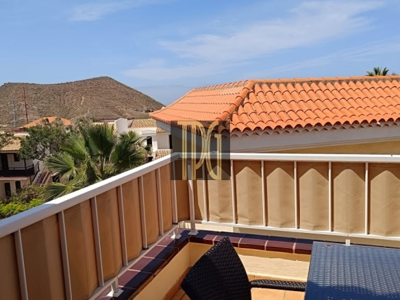 Terrace viewpoint with beige railing, wicker chair, and orange-tiled roof, overlooking palm trees and a rocky hill under a blue sky.