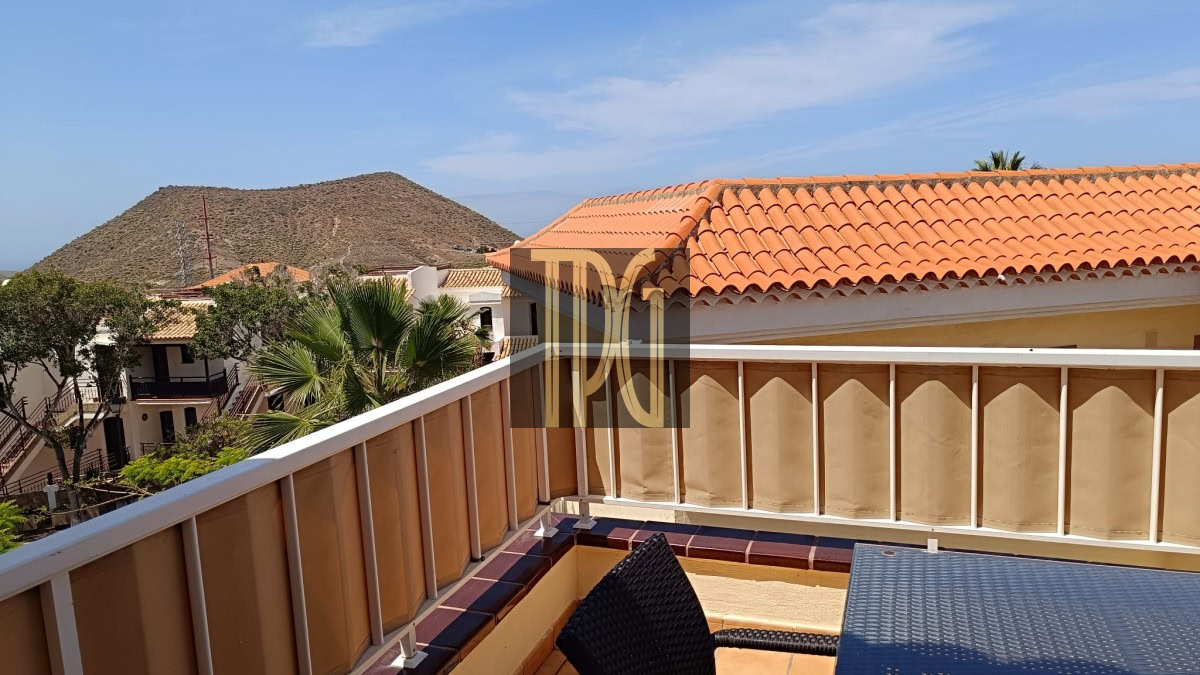 Terrace viewpoint with beige railing, wicker chair, and orange-tiled roof, overlooking palm trees and a rocky hill under a blue sky.