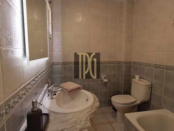 Small bathroom with a marble countertop sink, wall mirror, dark pump soap dispenser, beige hand towel, toilet, and a bathtub along tiled walls.