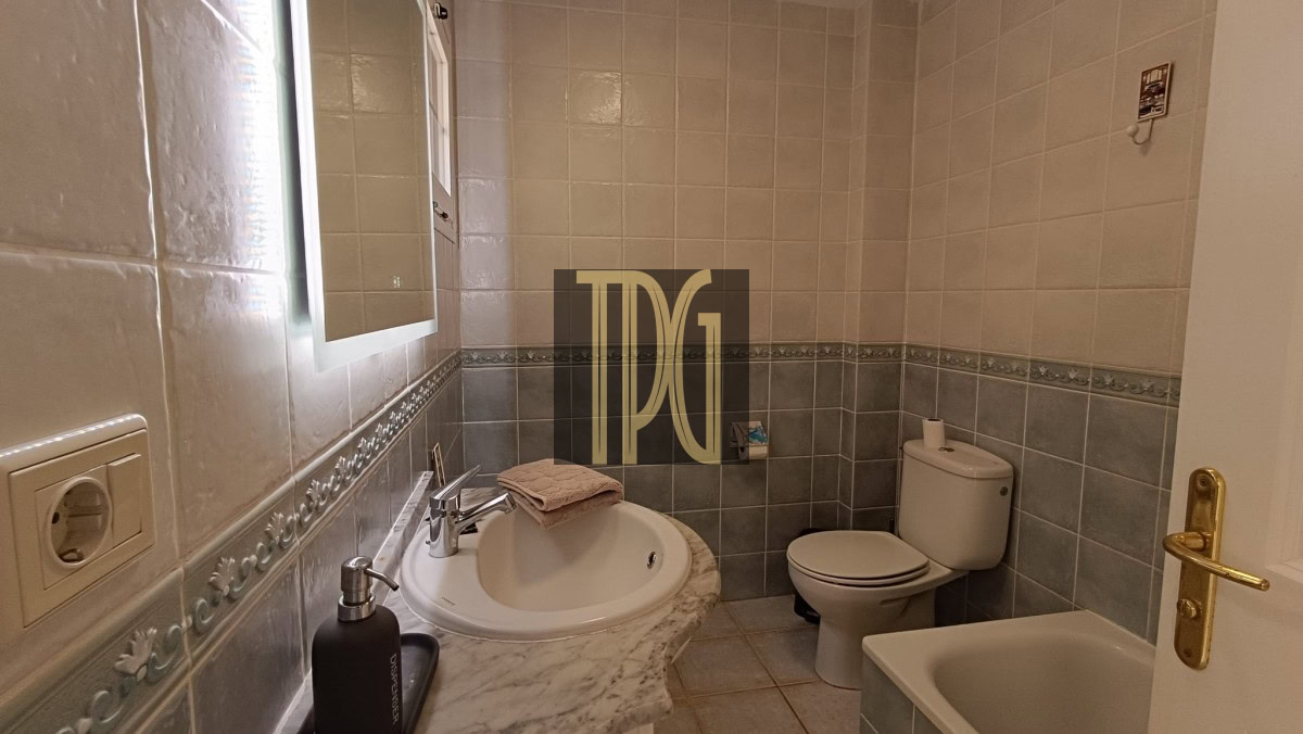 Small bathroom with a marble countertop sink, wall mirror, dark pump soap dispenser, beige hand towel, toilet, and a bathtub along tiled walls.