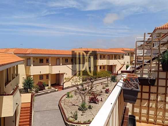 Sunny Mediterranean-style apartment complex courtyard with beige walls, orange-tile roofs, and a central garden under a blue sky.