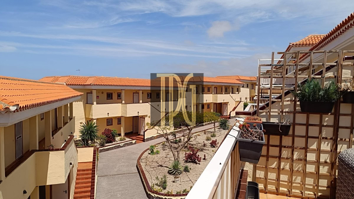 Sunny Mediterranean-style apartment complex courtyard with beige walls, orange-tile roofs, and a central garden under a blue sky.