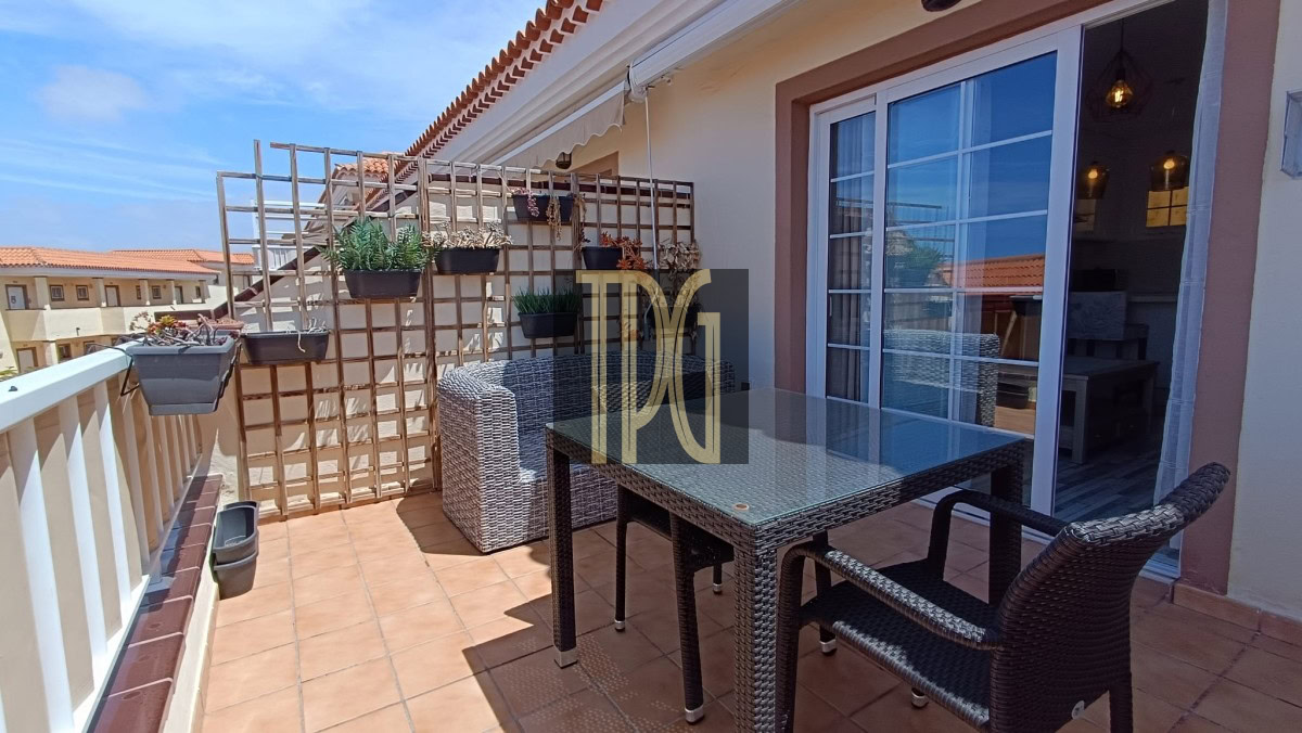 Outdoor balcony with a glass-top wicker table and chairs, potted plants on a trellis against a sliding door. I"m the text