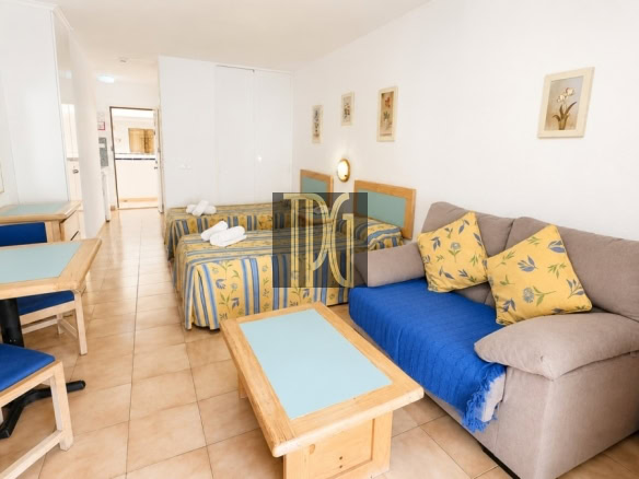 Living area of a compact hotel suite with a gray sofa, blue throw, yellow floral cushions, two twin beds, and a wooden coffee table.