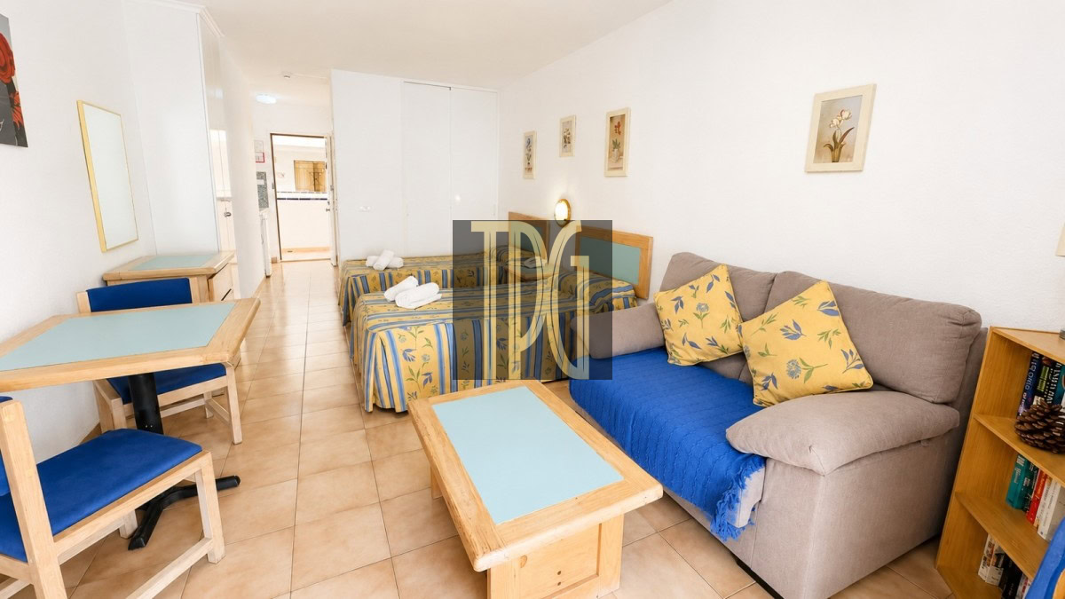 Living area of a compact hotel suite with a gray sofa, blue throw, yellow floral cushions, two twin beds, and a wooden coffee table.