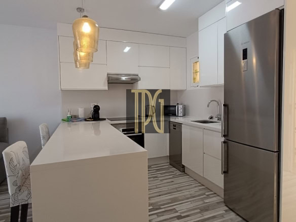 Modern kitchen with white cabinetry, an island, and a stainless steel fridge; living area with a large wall clock on the left.