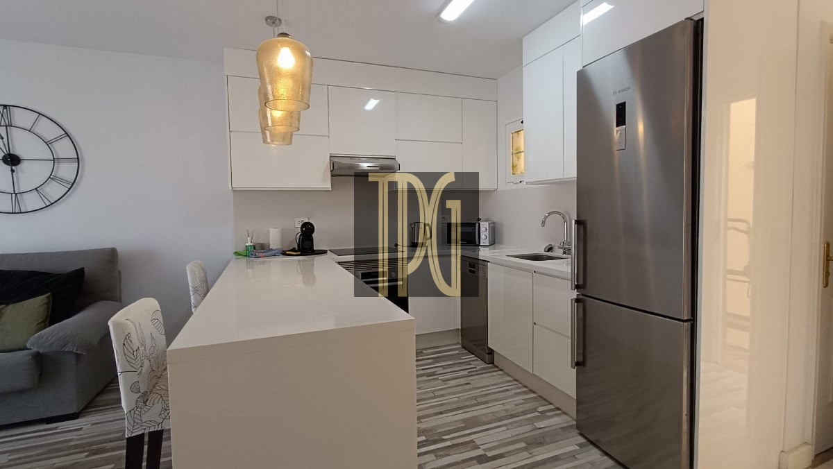 Modern kitchen with white cabinetry, an island, and a stainless steel fridge; living area with a large wall clock on the left.