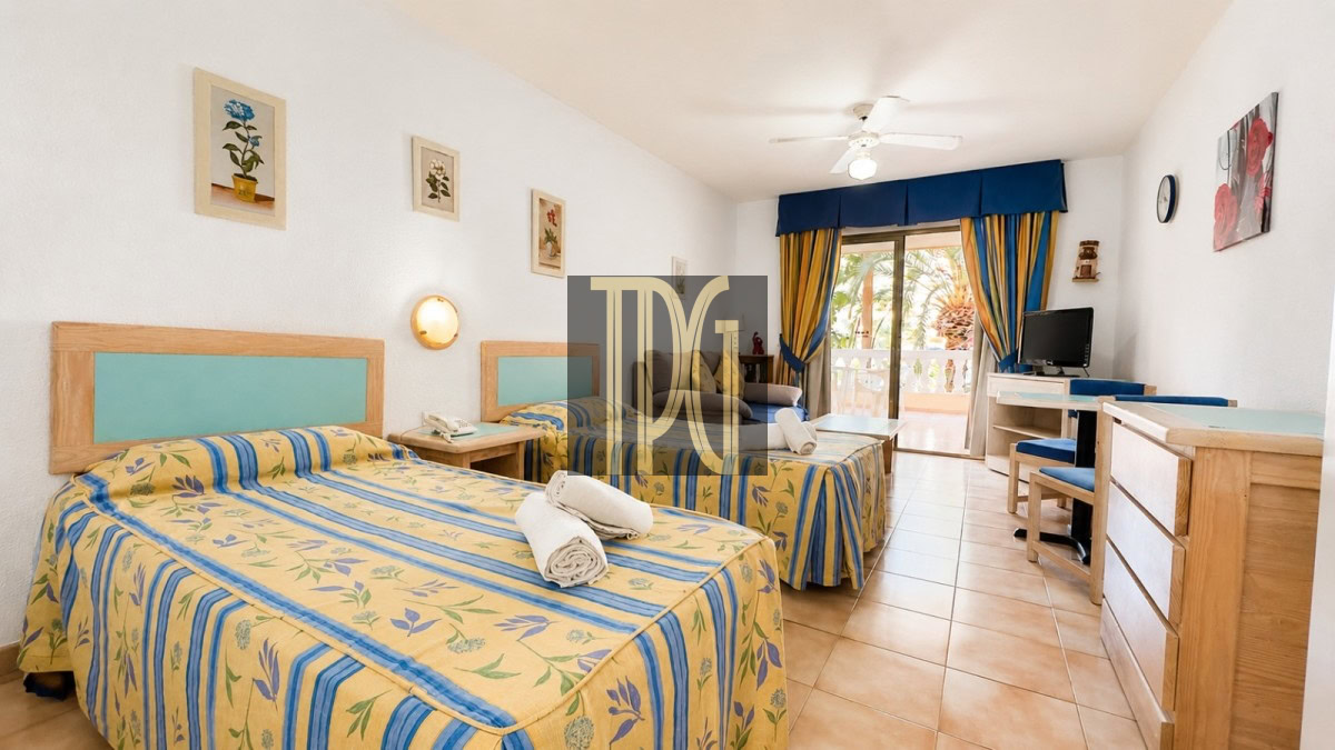 Twin-bed hotel room with yellow-blue floral bedspreads and turquoise headboards; towels on beds and a balcony door with blue curtains in background.