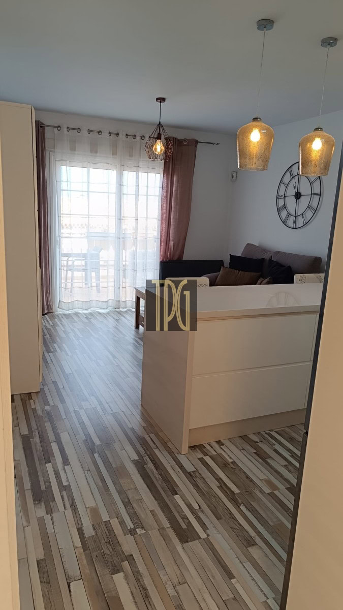 Bright open-plan living room with a white partition, gray sofa with cushions, and a large black wall clock; two pendant lights hang from the ceiling, and a glass door with sheer curtains opens to a balcony/ patio area.