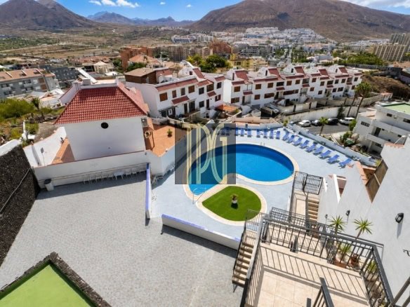 Aerial view of a resort's circular blue pool surrounded by lounge chairs, white buildings with red-tiled roofs, and distant mountains.