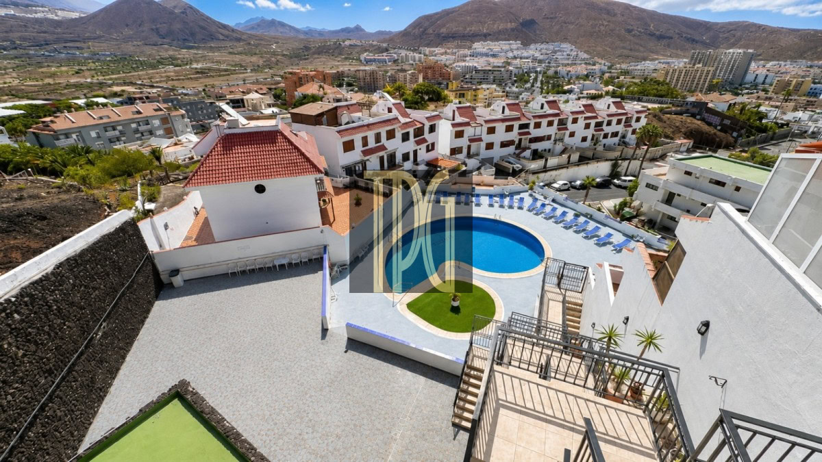 Aerial view of a resort's circular blue pool surrounded by lounge chairs, white buildings with red-tiled roofs, and distant mountains.