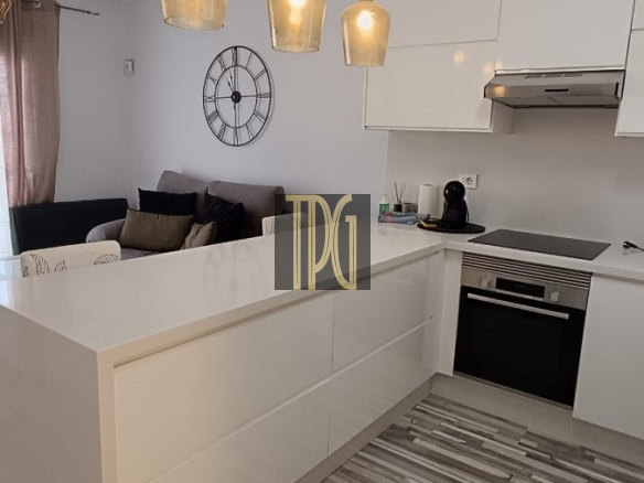 Open-plan kitchen with white island, pendant lights, and built-in oven; living room with clock on wall in background.