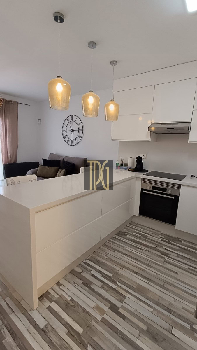 Open-plan kitchen with white island, pendant lights, and built-in oven; living room with clock on wall in background.