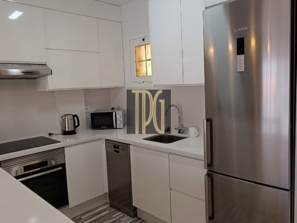 Modern white L-shaped kitchen with stainless steel refrigerator, oven, and dishwasher; grey striped floor and bright overhead light.