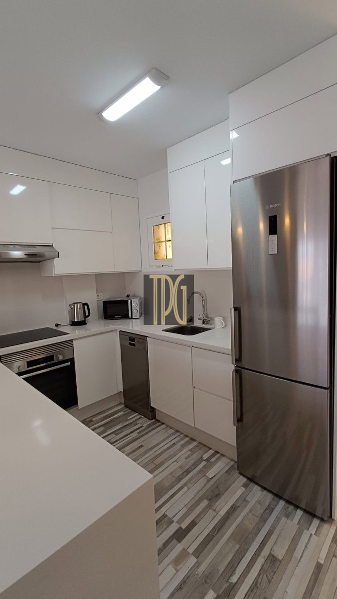 Modern white L-shaped kitchen with stainless steel refrigerator, oven, and dishwasher; grey striped floor and bright overhead light.