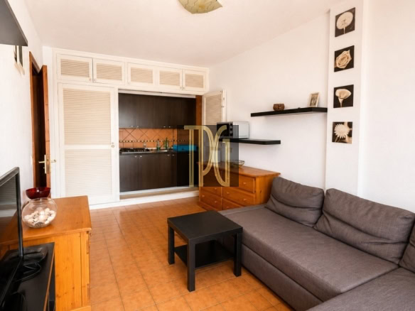 Living room with a gray sectional sofa, black coffee table, and TV on a wooden stand; open kitchen visible in the back.