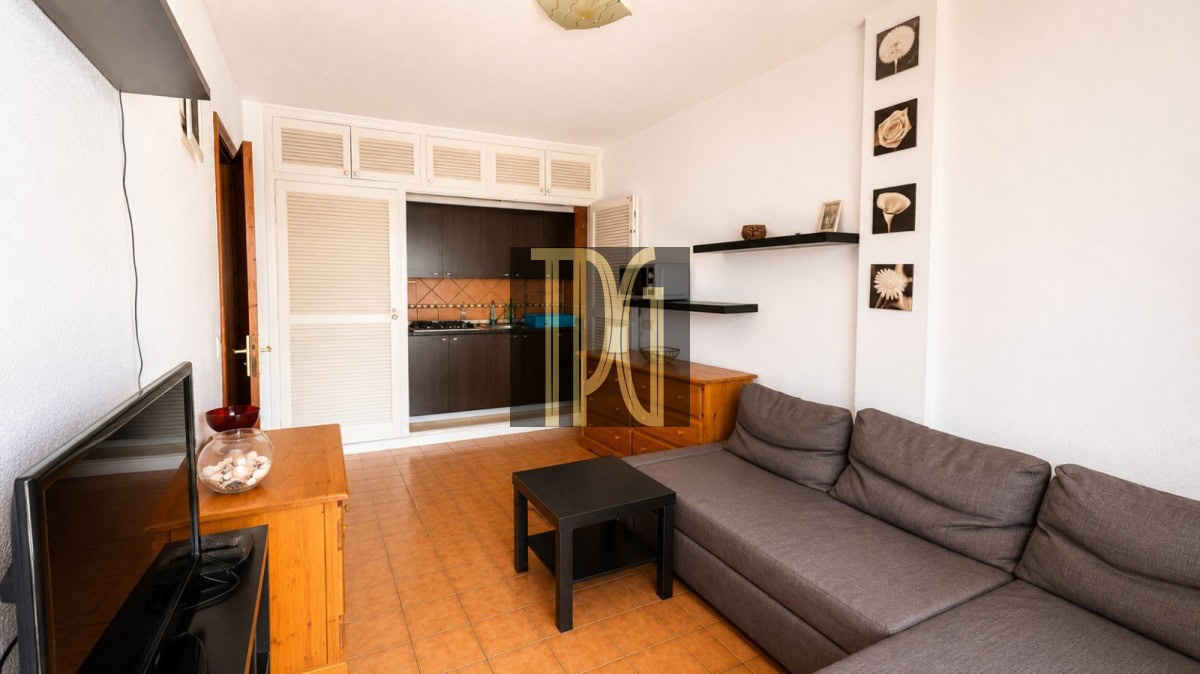 Living room with a gray sectional sofa, black coffee table, and TV on a wooden stand; open kitchen visible in the back.