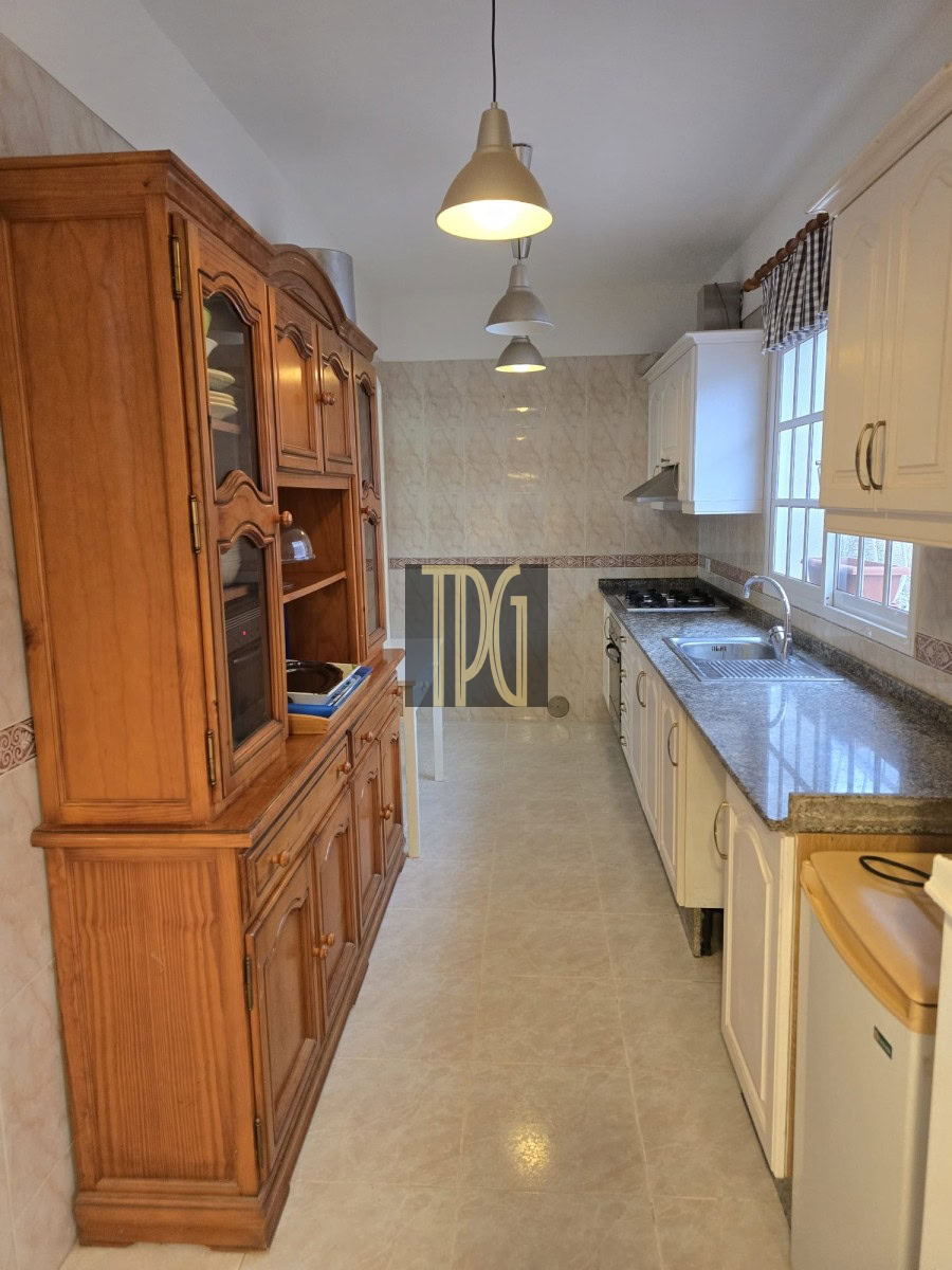 Narrow galley kitchen with a tall wooden cabinet on the left, white upper cabinets on the right, granite countertops, and three pendant lights overhead.